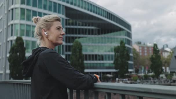 Sportliche Frau mit Kopfhörern steht auf Brücke des Stadtflusses — Stockvideo