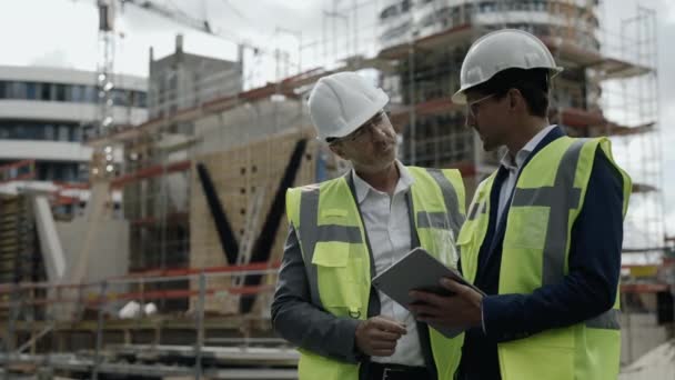 Dwóch budowniczych w kaskach korzystających z tabletu na placu budowy — Wideo stockowe