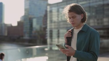 Sıradan giyinen bir adam nehir kenarında yürüyor ve cep telefonu kullanıyor.