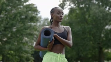 Afrikalı Amerikalı kadın parkta yoga minderiyle poz veriyor.
