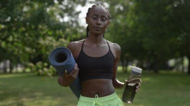 Saç örgülü, su şişesi ve yoga minderi taşıyan sportif bir kadın.