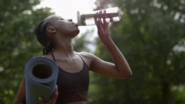 Yoga minderi tutan ve parkta su içen kadın.
