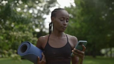 Cep telefonu kullanan ve yoga minderi taşıyan Afrikalı Amerikalı bir kadın.