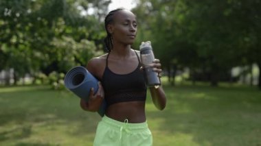 Spor kıyafetli, su içen ve yoga minderi taşıyan bir kadın.