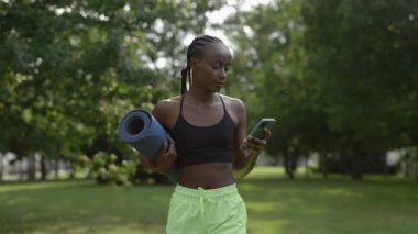 Spor kıyafetli bir kadın parkta cep telefonu ve yoga minderiyle yürüyor.