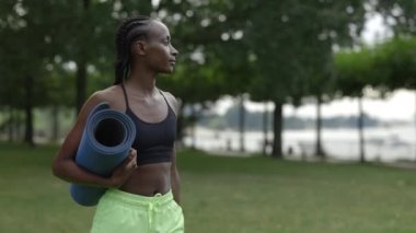 Yoga minderi ve bir şişe suyla parkta yürüyen bir kadın.