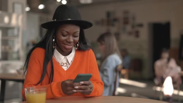 Glada afrikanska kvinna som har goda nyheter på smartphone när du sitter vid cafébordet. Söt kvinna i snygg outfit med mobiltelefon inomhus. — Stockvideo