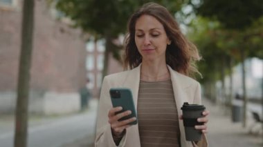 Meşgul bir kadın cep telefonundan mesaj atıyor ve dışarıda kahve taşıyor.