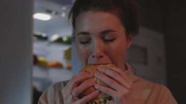 Woman eating burger at night near refrigerator
