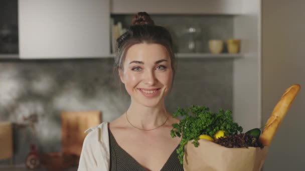 Uśmiechnięta kobieta trzyma torbę spożywczą stojąc na kuchni — Wideo stockowe