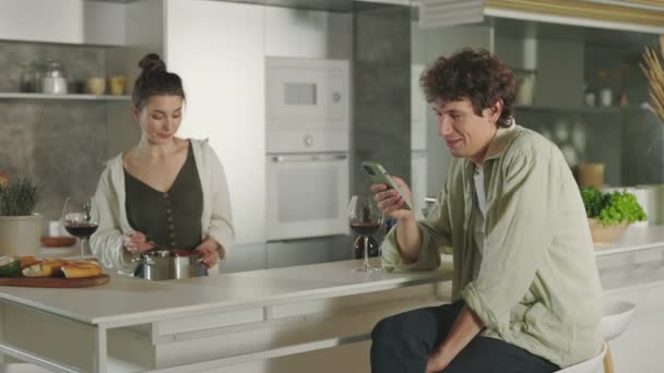 Mężczyzna za pomocą telefonu komórkowego i picia wina podczas gotowania kobieta — Wideo stockowe