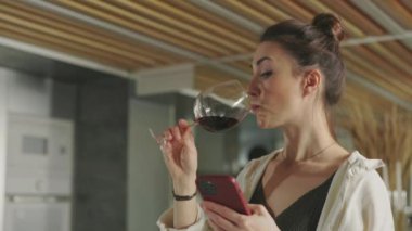 Kadın şarap içiyor ve adam yemek yaparken cep telefonu kullanıyor.