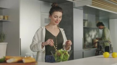 Kadın, kocası arka planda salata pişiriyor.