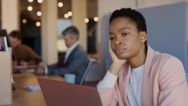 Mujer africana cansada sentada en la mesa y aburrida mientras trabaja apenas. Jovencita sintiéndose soñolienta durante el trabajo en el portátil. Concepto de exceso de trabajo. — Vídeos de Stock