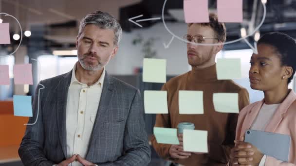 Trabajador de oficina masculino poniendo pegatinas en tablero de cristal y explicando el concepto a los miembros del equipo. Primer plano del hombre barbudo guapo de pie cerca de la pared de vidrio en la conferencia corporativa. — Vídeos de Stock