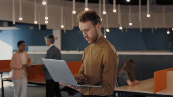 Erfolgreicher Geschäftsmann in formeller Kleidung, der mit Laptop in der Hand im Büro steht. Kaukasier lächelt in die Kamera. Geschäftsleute und Gadgets-Konzept. — Stockvideo