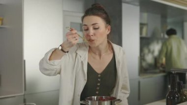 Kadın yemeğin tadına bakarken, erkek arka planda yemek yapıyor.