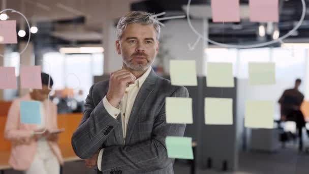 Vážný obchodní manažer zkoumá informace o lepkavých poznámkách a nalézá řešení. Muž korporátní lídr při pohledu na skleněnou stěnu se samolepkami v kanceláři. — Stock video