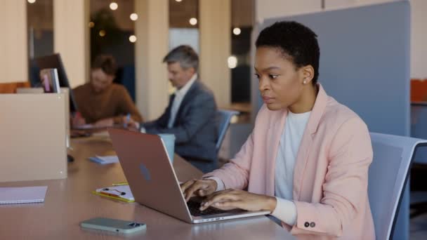 Joven trabajadora de oficina multirracial regocijándose de algo mientras mira su portátil moderno. Chica africana sentada en la oficina y trabajando. — Vídeos de Stock