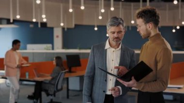 Beyaz, genç bir adam ve kıdemli bir adam ofis merkezinde durmuş gülümsüyor ve konuşuyorlar. Belge ve el sıkışma ile klasörü tutan işadamları.