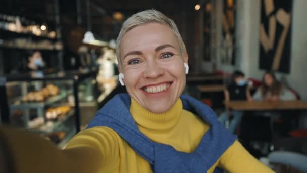 ผู้หญิงยิ้มและโบกมือระหว่างการโทรวิดีโอที่ร้านกาแฟที่อบอุ่น — วีดีโอสต็อก