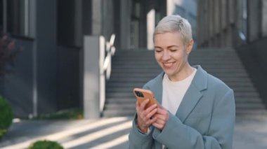 İş merkezinin yakınında yürürken akıllı telefon kullanan mutlu orta yaşlı beyaz kadın. Teknoloji, iletişim, iş konsepti.