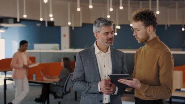 Dois parceiros de negócios do sexo masculino trabalhando juntos em pé no centro de negócios e discutindo projeto comum usando tablet digital. Comunicação, conceito de negócio. — Vídeo de Stock