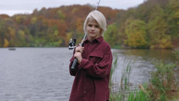 Caucasian boy with fishing rod on shoulder posing near lake — Stock Video