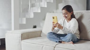 Akıllı telefonuyla kanepede otururken gülümseyen genç bir kız.