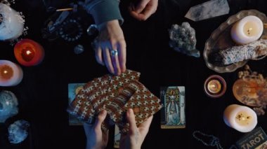 New York, ABD - 1 Mayıs 2021: Falcı masadaki tarot kartlarını ayarlıyor ve müşteri karanlık odada bunlardan birini çekerken elinde fanı tutuyor.