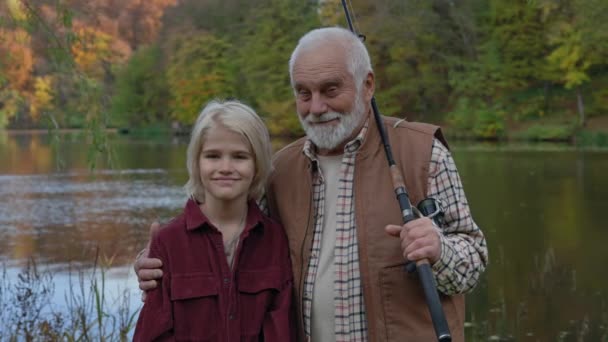 Porträtt av lycklig åldrad man kramar sitt barnbarn tittar på kameran står tillsammans nära sjön. Kaukasiska familjen fiske på lokal sjö. — Stockvideo