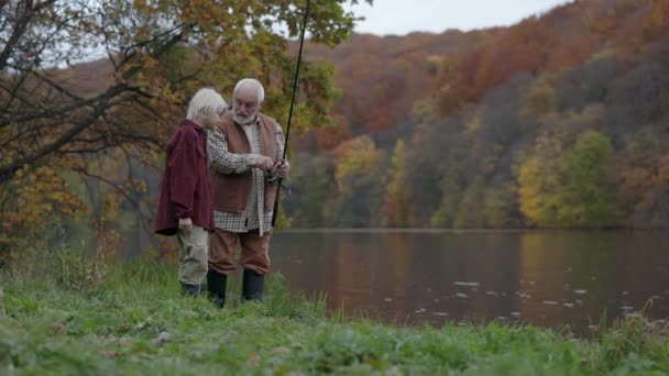Farfar med fiskespö pratar med sonson nära sjön — Stockvideo