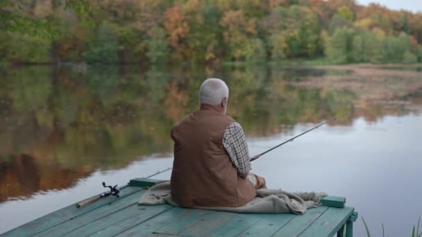 Rückansicht eines alten Mannes, der auf einem hölzernen Steg sitzt und fischt — Stockvideo