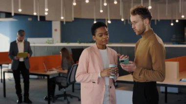 Kendine güvenen bir ofis çalışanı akıllı telefondaki bayan iş arkadaşına bir şey gösteriyor. İş arkadaşları ellerinde kahve fincanlarıyla salonda durup ekrana bakıyorlar. İnsanlar ve teknoloji kavramı.