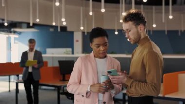 Akıllı telefondaki bayan iş arkadaşına bir şey gösteren yakışıklı bir ofis çalışanı. İş arkadaşları ellerinde kahve fincanlarıyla salonda durup ekrana bakıyorlar. İnsanlar ve teknoloji kavramı.