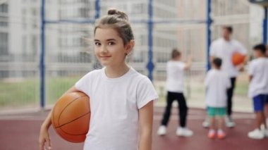 Small beautiful girl standing with ball outdoors in school basketball court. Portrait female school children standing on the sport yard background. Sport and education concept