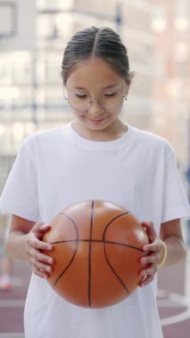 Vertical video. Poprtrait asian schoolgirl standing on basketball court holding ball raise head and smiling on the camera. Sport, education and people concept. Close up