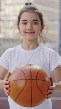 Vertical video. Portrait of smilling schoolgirl standing on court holding ball looking at camera. Sport, education and people concept. Close up