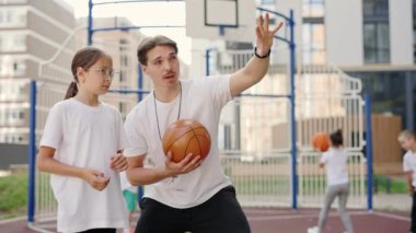 Handsome male coach explaining the throwing technique of basketball game. Team of children play a game with ball on the background. People, sport and education concept.