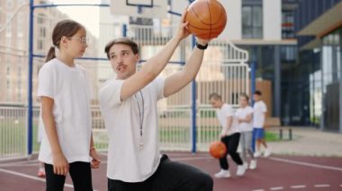 Handsome male coach explaining to child how to put a ball into the basket on court near school. Education and sport concept