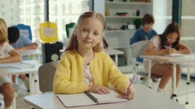 Portrait of a beautiful blonde pupil schoolgirl sitting on a chair in the classroom against the background of children holding a pen to write something down. People and educational concept