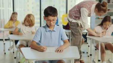Asian boy pupil wearing glasses sits at a first desk in the classroom and fills out tests, and the teacher in the background help students with their test. People and educational concept