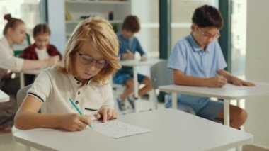 Red-head pupil wearing glasses sits at a first desk in the classroom and fills out tests thinking and writing down answers. People and educational concept