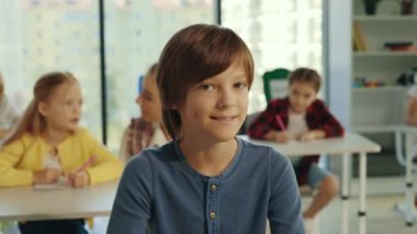 Portred of male smiled boy pupil looking at the camera sitting on the chair in the classroom and other pupils. People and educational concept
