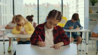 Beautiful long-haired brunette asian pupil wearing glasses sits at a first desk in the classroom and fills out tests thinks about answers, and other pupils doing tests on the background
