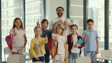 Portrait of the children stand with the male smiled teacher in the middle of the class and show a thumb up feeling happy and cheerful together in the private school. People and educational concept