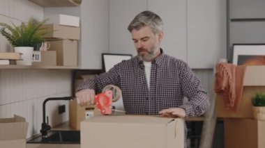 Man is taping a box in home. Middle aged man is moving and packing dishes on the kitchen background. New home and moving concept