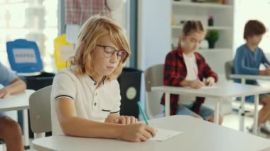 Red-head pupil wearing glasses sits at a desk at school and fills out tests, and the teacher in the background gives an assignment. People and educational concept