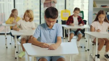 Asian boy pupil wearing glasses sits at a first desk in the classroom and fills out tests thinks about answers, and the teacher standing in the background. People and educational concept