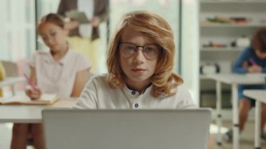 Portrait of the red-head boy sitting on the desk with laptop in a class during computer science lesson looking at the camera and other pupils and the teacher on the background